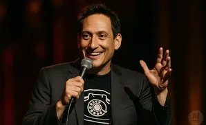 Comedian laughs into a microphone, wearing a black suit and a t-shirt with a rotary phone design.