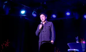 Comedian Bob Saget performs on stage, holding a microphone and wearing a dark shirt and hat.