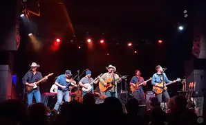 A band plays on a stage with red and white lights, instruments include guitars, banjo, and violin.