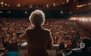 Person with white hair at podium addresses a large, blurred audience in a theater.