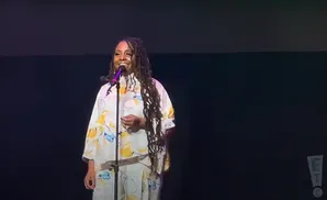 A woman with dreadlocks stands on stage, speaking into a microphone. She wears a white outfit with a floral pattern.