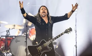 A man with long hair and a beard sings with his arms outstretched while playing a black electric guitar.