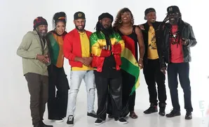 A group of seven people, dressed in Rastafarian colors, pose for a photo against a white backdrop.