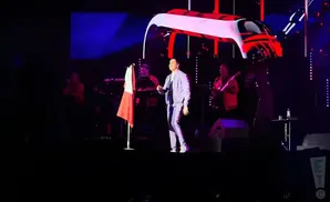 A performer in a light blue suit stands on stage with a band, holding a flag.