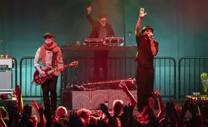 Musicians perform on stage with a crowd cheering in the foreground.