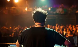 A DJ from behind, facing a blurred crowd at a concert, with stage lights above.