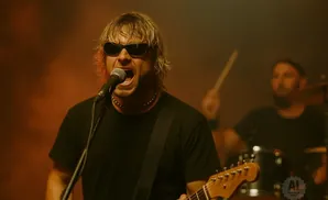 A man with sunglasses sings into a microphone while playing a guitar on stage.