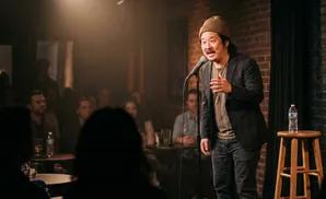 A comedian in a beanie and jacket performs on stage in front of a seated audience.