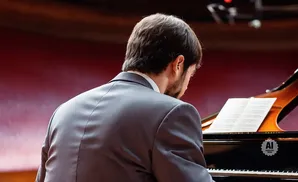 Man in a suit playing a grand piano, viewed from behind.