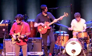 A band plays music on stage, with a guitarist in red, a bassist in a dark shirt, and a drummer in a white shirt.