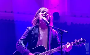 Musician with long hair and glasses plays an acoustic guitar and sings into a microphone on a purple-lit stage.