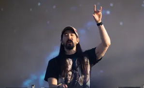 DJ Steve Aoki performs on stage, raising his hand in a rock-on gesture.