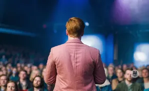 A person with red hair in a pink suit faces away from the camera, addressing a crowd in a dimly lit room with blue stage lighting.