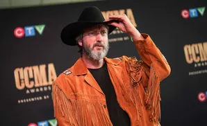 A man in a cowboy hat and fringed jacket adjusts his hat, posing in front of a black backdrop with logos.