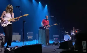 A band plays on a stage with blue spotlights. The bassist is in the foreground.