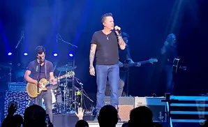Singer performing on stage with a band, wearing a black t-shirt and jeans, holding a microphone.