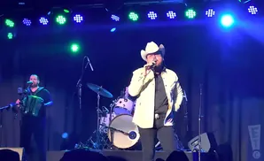 A male singer in a cowboy hat sings into a microphone on stage with a band.