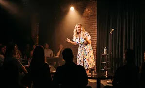 A female comedian in a colorful dress performs for an audience in a dimly lit club.