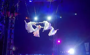 Four aerialists in white dresses perform a synchronized routine suspended from the ceiling.