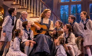Woman plays guitar and sings with a group of children in sailor-style outfits.