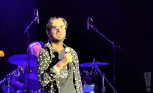 Musician Ringo Starr on stage, holding a microphone and wearing a black shirt with white stars and moons.