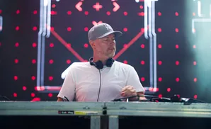 DJ with headphones around neck, wearing a grey cap and white t-shirt, stands behind a DJ console with a blurred stage background.