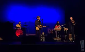 A band performs on a dimly lit stage with a bright blue backdrop. A guitarist sings into a microphone, while a violinist plays.