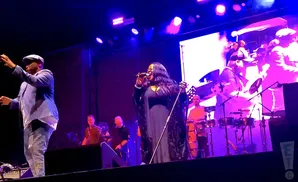 A male singer in a light blue suit and cap gestures to the left as a female singer in a black gown sings into a microphone.