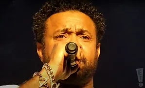 A close-up of a man with curly hair singing into a microphone, wearing gold bracelets with charms.