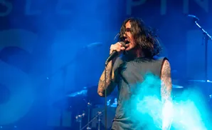 A male singer with long, dark hair sings into a microphone on a stage illuminated by blue lights and smoke.