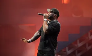 Rapper 50 Cent performs on stage wearing a black t-shirt, black baseball cap, and jeweled pants.
