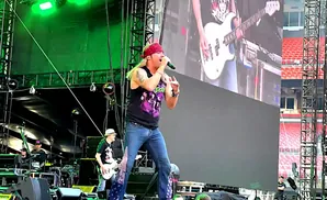 A male singer in a bandana and tank top sings into a microphone on a concert stage with a large screen behind him.