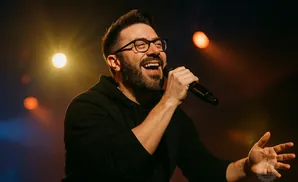 Man with beard and glasses singing into a microphone on a dimly lit stage.