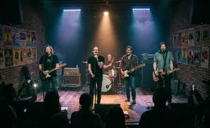 A band performs on a brick stage lit by spotlights, with posters on the walls and an audience in the foreground.