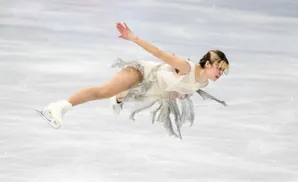 A figure skater in a light-colored dress leaps in the air, her arms outstretched.