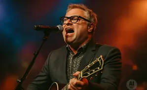 A man with glasses sings into a microphone while playing a guitar.