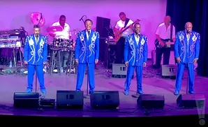 Five men in matching blue suits with silver accents perform on stage, with band members playing drums, bass, and keyboards behind them.