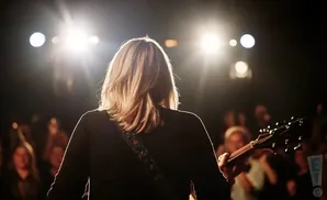 A musician with blonde hair plays guitar on a dimly lit stage with bright spotlights and an audience in the background.