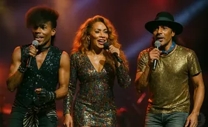 Three singers on stage: a man in a sequined vest, a woman in a sequined dress, and a man in a sequined shirt and hat.