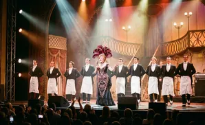 A singer in a feathered headdress performs on stage with a chorus of men in tuxedos.