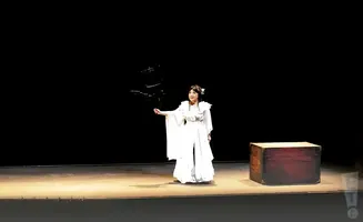 A woman in a white gown stands on a stage, holding a prop. A wooden chest sits to her right.