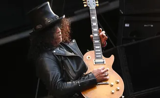 A guitarist with curly hair wearing a top hat and leather jacket plays a Gibson Les Paul guitar.