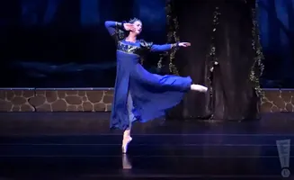 A ballerina in a blue dress and crown performs a graceful pose on stage.
