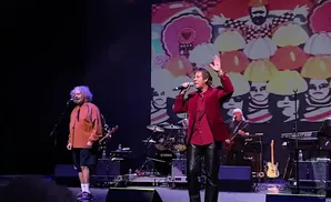 A male singer in a red jacket and leather pants performs on stage with a band behind him.