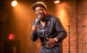 A Black man in a cap and dark shirt laughs while holding a microphone on stage.