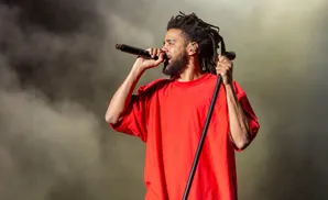 Rapper J. Cole performs on stage, wearing a red shirt and holding a microphone.
