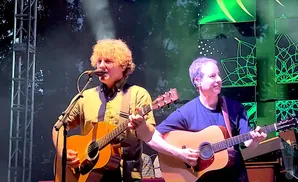 Two men playing acoustic guitars on stage with a green, patterned backdrop.