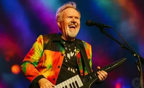 A man with white hair and a beard laughs while playing a keytar on a colorful stage.