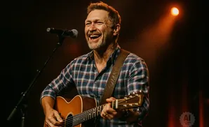 A man in a plaid shirt laughs while playing an acoustic guitar and singing into a microphone on stage.