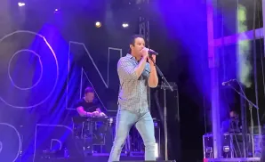 A male singer in a plaid shirt and jeans performs on a stage with a large banner and blue lighting.